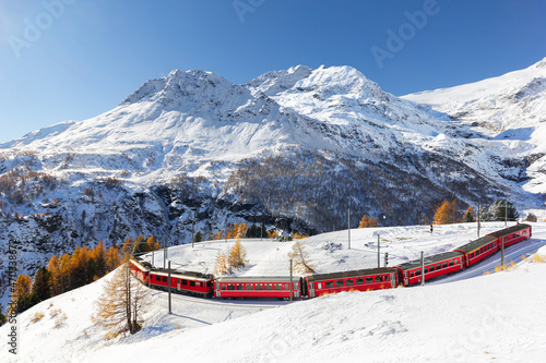A red train is passing the train tracks with tight 180° curve at high Alp Grum. The Piz Palu peak is at the background. photo