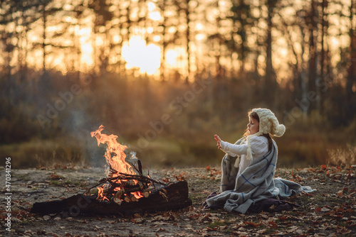 A child, a girl warms her hands by a campfire in the forest, wrapped in a blanket.