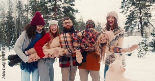 Fun happy multiethnic friends hold, rock young blonde woman laughing at snowy forest Christmas vacation slow motion.