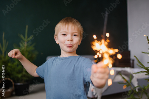 The boy toddlets a sparkler. Christmas mood holiday atmosphere. Home fireworks. Lifestyle childhood moments. The boy is blond. Green background. Cozy bedroom interior. Having fun. Enjoys shows tongue © Antipina