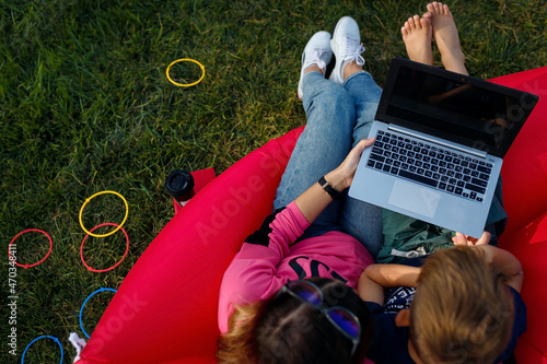 Mother with laptop, her barefoot son sitting on red Lamzac Hangout outdoors on green grass lawn, top view. Leisure time and work with child photo
