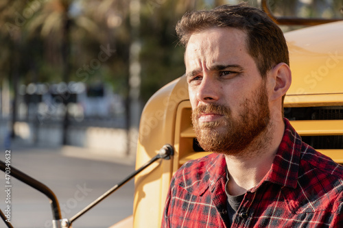 Young man in retro style and red plaid shirt staring leaning on the hood of a yellow bus