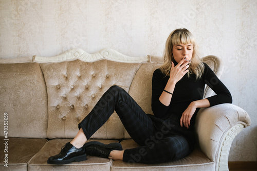 Fashion portrait of beautiful blond young woman, she is sitting on the sofa and smoking a cigarette