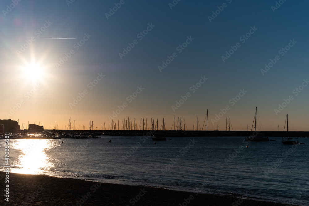Sun flare shining on a blue solid morning sky on a quiet sea shore landscape with boat silhouettes in a domestic dock