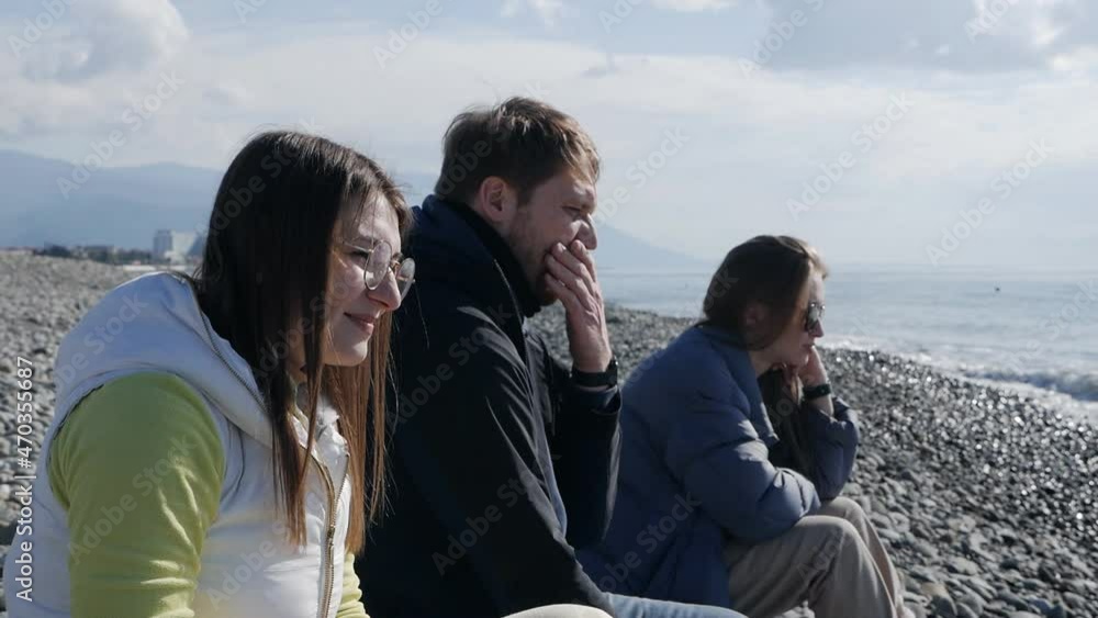 Friends are sitting on the shore of a stone beach by the sea. In the cool autumn, a man and two girls enjoy the sea air. Throw stones into the water into the sea.Slow motion Friends are sitting on the shore of a stone beach by the sea. In the cool autumn, a man and two girls enjoy the sea air. Throw stones into the water into the sea.Slow motion