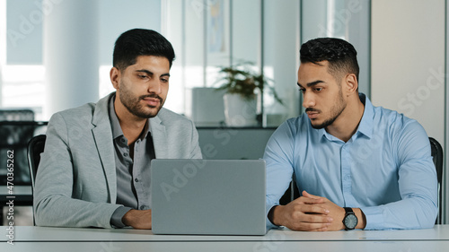 Two serious focused Arab young male businessmen sitting in modern office using laptop looking at computer screen. Diverse employees East Indian colleagues discussing working together checking emails