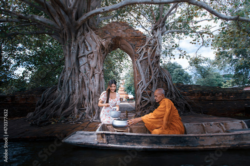 Fotografia Young woman give alms to a Buddhist monk at front of ancient gate of Pra Ngam temple under Bodhi Tree during monk transport via wooden boat when Ayutthaya flooded at Phra Ngam temple of Thailand