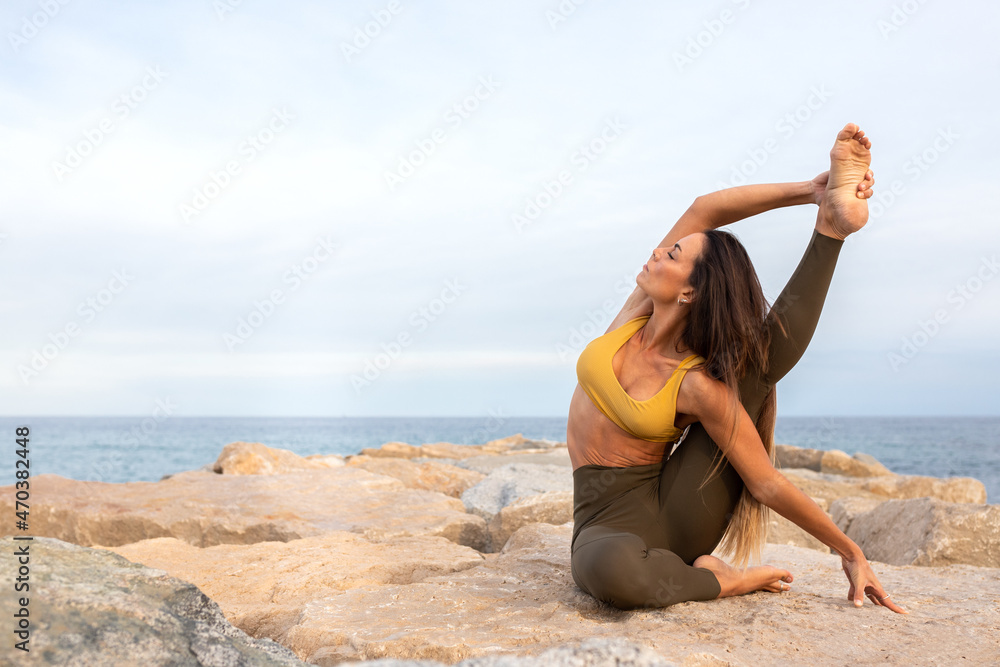 Flexible young woman doing Parivritta Kraunchasana yoga pose on a rock near the sea. Copy space.