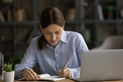 Concentrated serious young woman sit at desk near laptop jotting information in notebook. Making important notes, write personal records to daybook, keep a diary, create business to-do list concept
