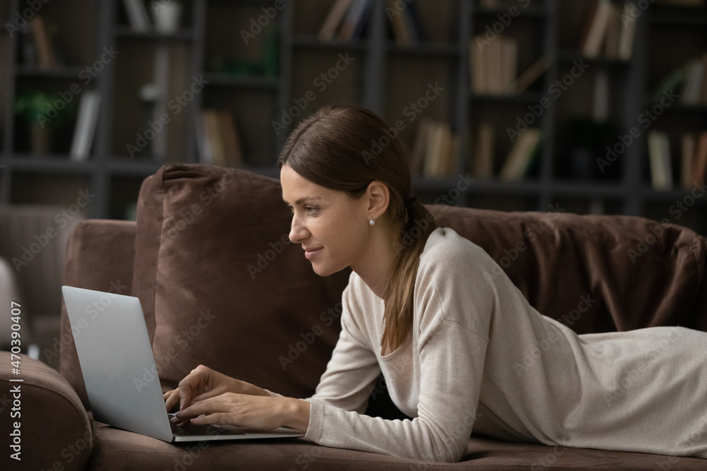 Young 30s woman lying on couch in living room working online using wireless computer. Female relax on sofa at home looks at laptop screen writing message, chat in social media. Communication concept Young 30s woman lying on couch in living room working online using wireless computer. Female relax on sofa at home looks at laptop screen writing message, chat in social media. Communication concept