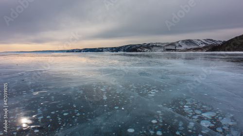 Sunset over a frozen lake. Cracks and methane bubbles are visible on the smooth, shiny ice. Sun glare on the surface. Mountains against the background of a pinkening cloudy sky. Baikal