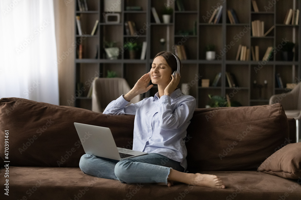 Serene woman relaxing alone on sofa in living room with eyes closed listen song through wireless headphones using online music streaming services app on laptop. Modern tech, connection, hobby concept Serene woman relaxing alone on sofa in living room with eyes closed listen song through wireless headphones using online music streaming services app on laptop. Modern tech, connection, hobby concept