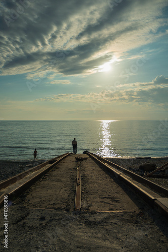 Fototapeta Naklejka Na Ścianę i Meble -  A man stands on a concrete pier going into the sea. On the water track from the evening sun. Blue sky with beautiful clouds. A woman goes swimming in the sea.