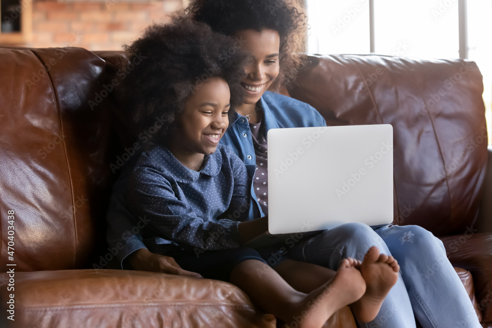 Caring young African American mother teaching little child daughter using computer apps, resting on couch at home. Addicted to modern technology bonding two generations diverse family playing online. Caring young African American mother teaching little child daughter using computer apps, resting on couch at home. Addicted to modern technology bonding two generations diverse family playing online.