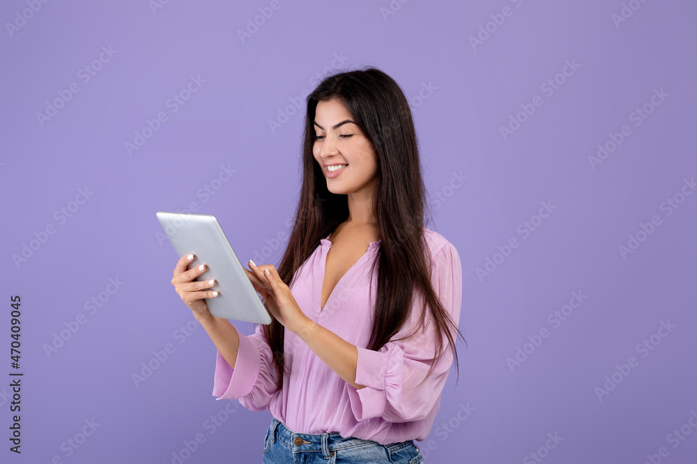Happy armenian lady using digital tablet, looking at screen and smiling, standing over purple background, copy space Happy armenian lady using digital tablet, looking at screen and smiling, standing over purple background, copy space