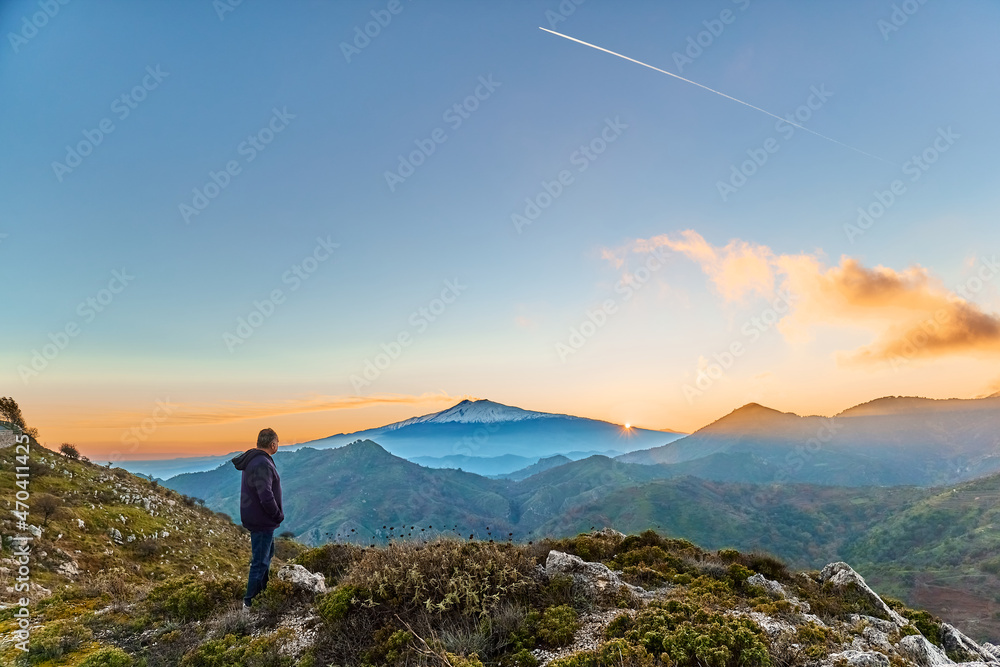 Man standing on top of the cliff in the mountains at sunset enjoying the beautiful sunset over erupting volcano Etna. Beautiful view in Sicily, Italy