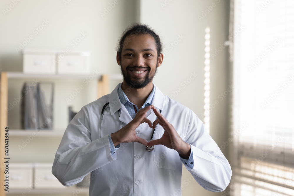 Obraz premium Head shot portrait smiling young African American man doctor in white uniform with stethoscope showing heart gesture, looking at camera, standing in hospital office, expressing support and care