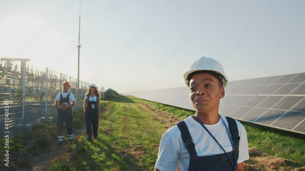 Professional young african smiling woman engineer wearing safety suit and helmet walking proudly on modern solar station. Sustainable plant. Clean energy development.