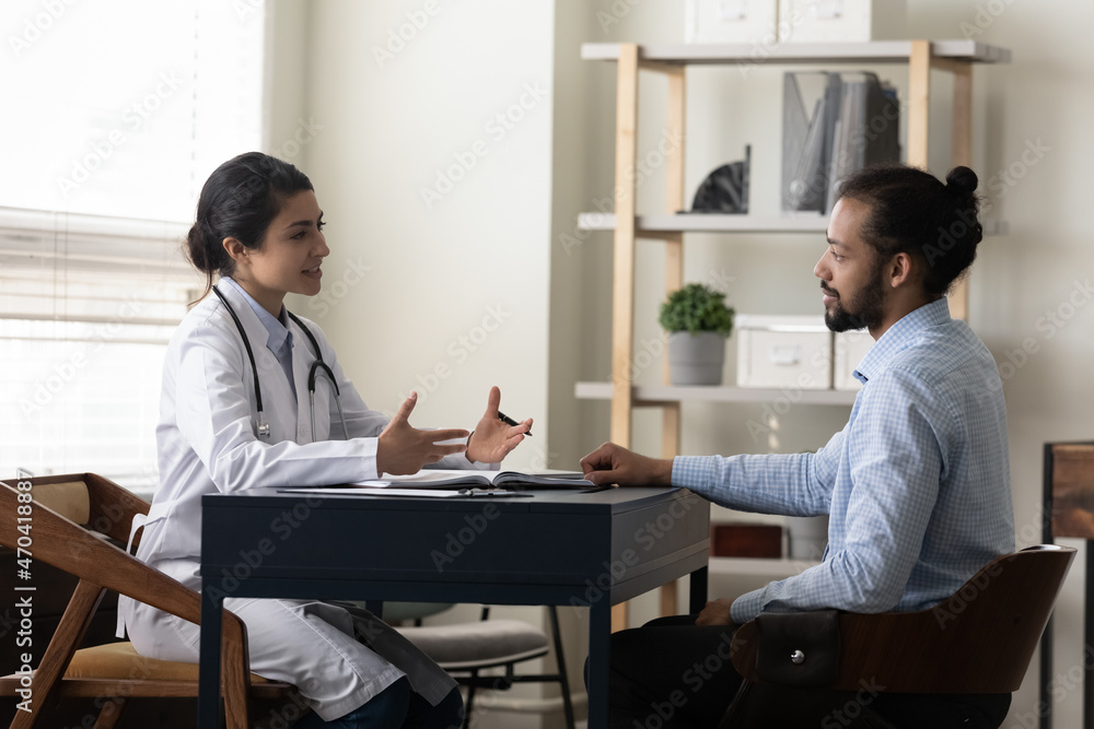 Smiling Indian woman doctor talking, consulting African American man patient at meeting in hospital, young female physician practitioner giving recommendation, discussing checkup or symptoms Smiling Indian woman doctor talking, consulting African American man patient at meeting in hospital, young female physician practitioner giving recommendation, discussing checkup or symptoms