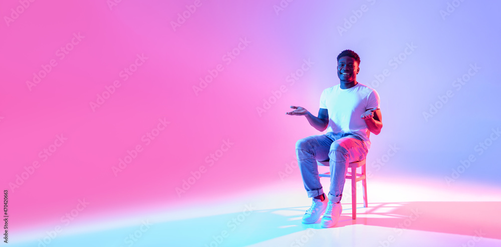 Full length of happy black guy sitting on chair, offering free space for your advertisement in neon light, banner Full length of happy black guy sitting on chair, offering free space for your advertisement in neon light, banner