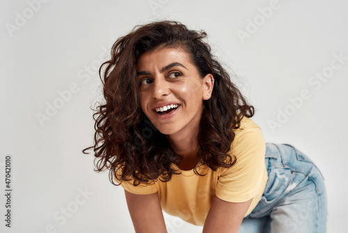 Happy young woman with dark curly hair in casual wear looking cheerful  smiling aside while posing isolated over gray background