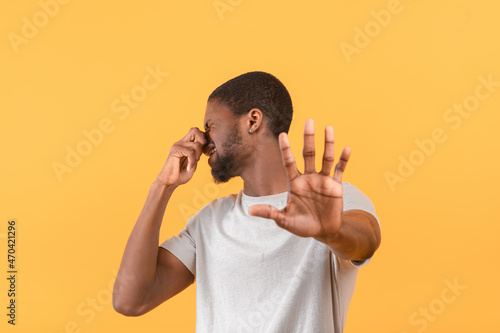 Terrible smell concept. Black guy closing nose and gesturing STOP  feeling disgusted  standing over yellow background