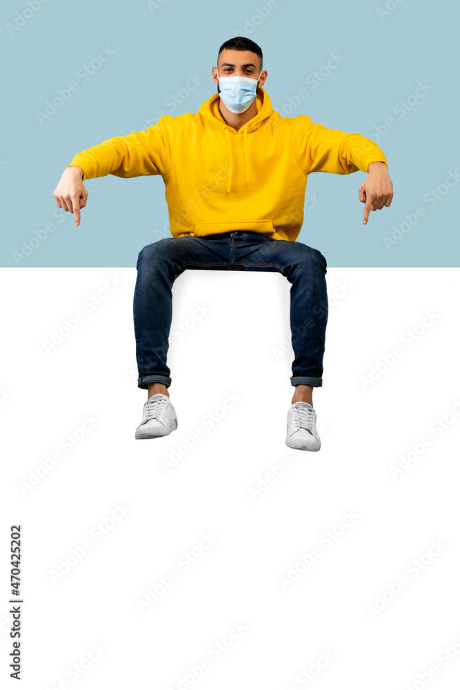 Young arab man in protective mask pointing at huge empty board for advertisement, sitting on it over blue background Young arab man in protective mask pointing at huge empty board for advertisement, sitting on it over blue background