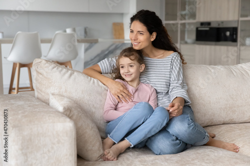 Smiling dreamy woman with cute little daughter kid visualizing good future, sitting relaxing on cozy couch at home, looking to aside, happy young mom with adorable girl child dreaming together