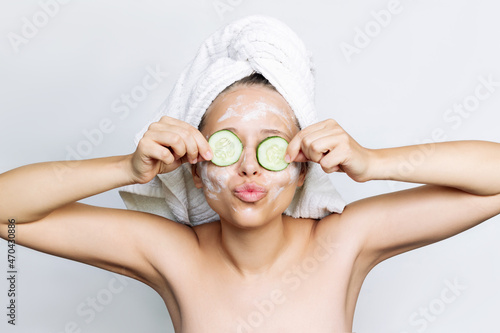 A young funny woman with a white towel on her head after a shower holds cucumber slices, covers her eyes making a face mask, sending an air kiss isolated on a gray background. Skin care, cosmetology