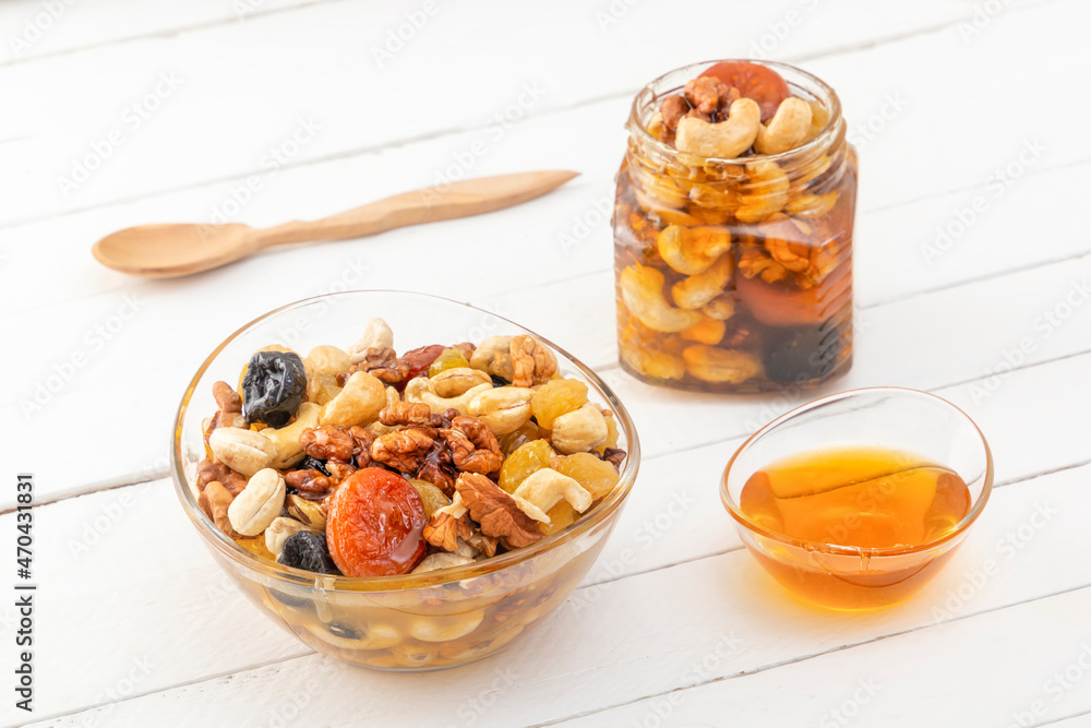 Glass cup and jar filled with a mixture of walnuts, cashews, raisins, dried apricots and prunes with honey all together with a wooden spoon on a white board table Glass cup and jar filled with a mixture of walnuts, cashews, raisins, dried apricots and prunes with honey all together with a wooden spoon on a white board table