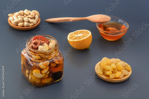 Mixture of walnuts, cashews, raisins, dried apricots and prunes with honey in a glass jar along with a spoon and lemon on a gray table