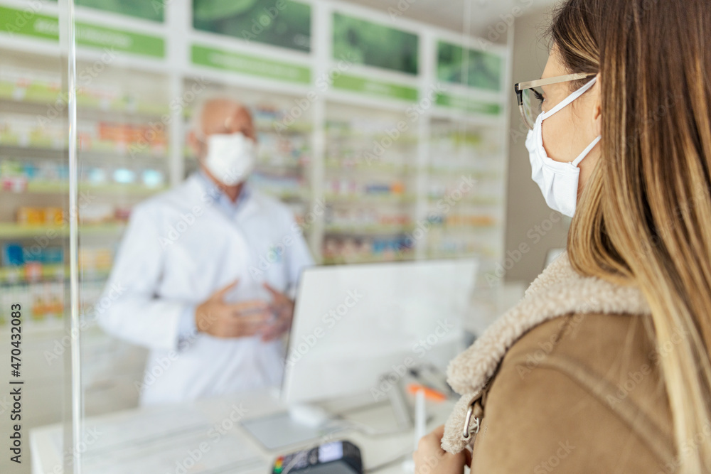 Buying and selling prescription drugs and pharmacist advice. A male pharmacist blurred in the background behind the counter selling drugs to an adult girl wearing a protective mask during corona virus Buying and selling prescription drugs and pharmacist advice. A male pharmacist blurred in the background behind the counter selling drugs to an adult girl wearing a protective mask during corona virus