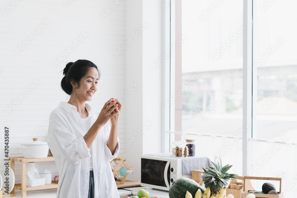 Asian woman holding apples in the kitchen. Woman eating fresh apple while standing in the kitchen.