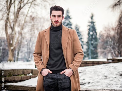 Young man in snowy city park wearing wool coat, looking at camera
