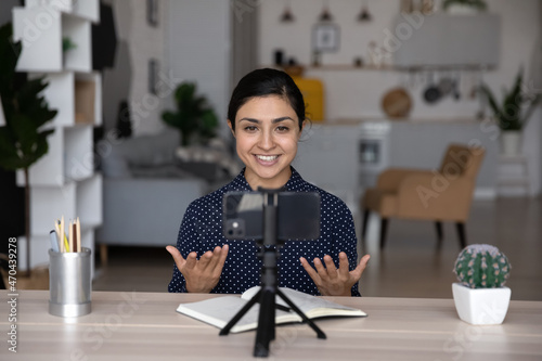 Confident smiling young Indian ethnicity female blogger recording educational video on cellphone standing on stabilizer, sharing professional knowledge, streaming live stories online, sitting at table