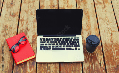 Close up laptop and book with eyeglasses, cup of coffee lying on wooden table or floor background, blank screen, top view