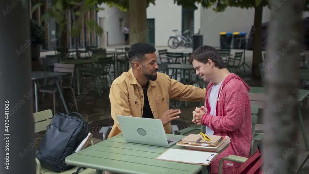 Young man with Down syndrome with his mentoring friend sitting outdoors in cafe and studying. Young man with Down syndrome with his mentoring friend sitting outdoors in cafe and studying.