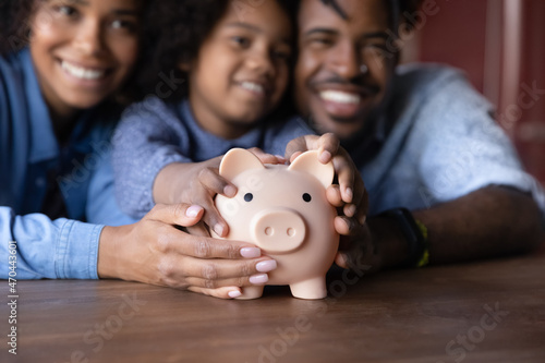 Joyful African family young couple parents and little child girl holding piggybank in hands, feeling excited of saving money, planning future expenditures, learning managing budget at home.