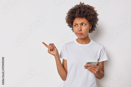 Upset discontent woman with curly hair points away with index finger shows blank copy space for your advertising uses smartphones dressed casually isolated over white background doesnt like something photo