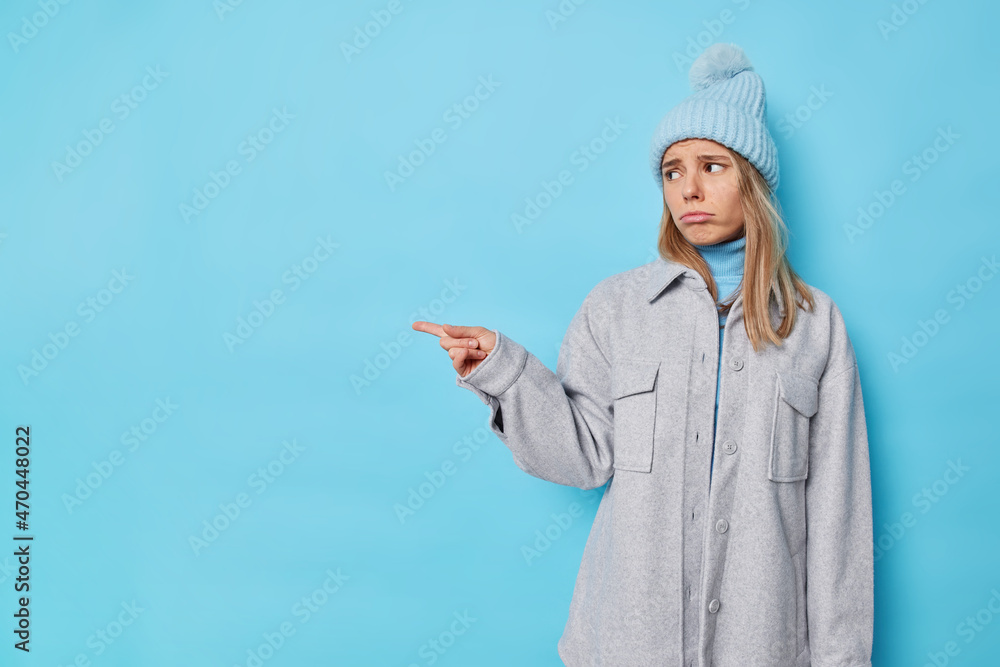 Upset disappointed woman points away with index finger dislikes product givesnegative feedback stands unhappy wears hat and grey jacket isolated over blue background has unsatisfied expression Upset disappointed woman points away with index finger dislikes product givesnegative feedback stands unhappy wears hat and grey jacket isolated over blue background has unsatisfied expression