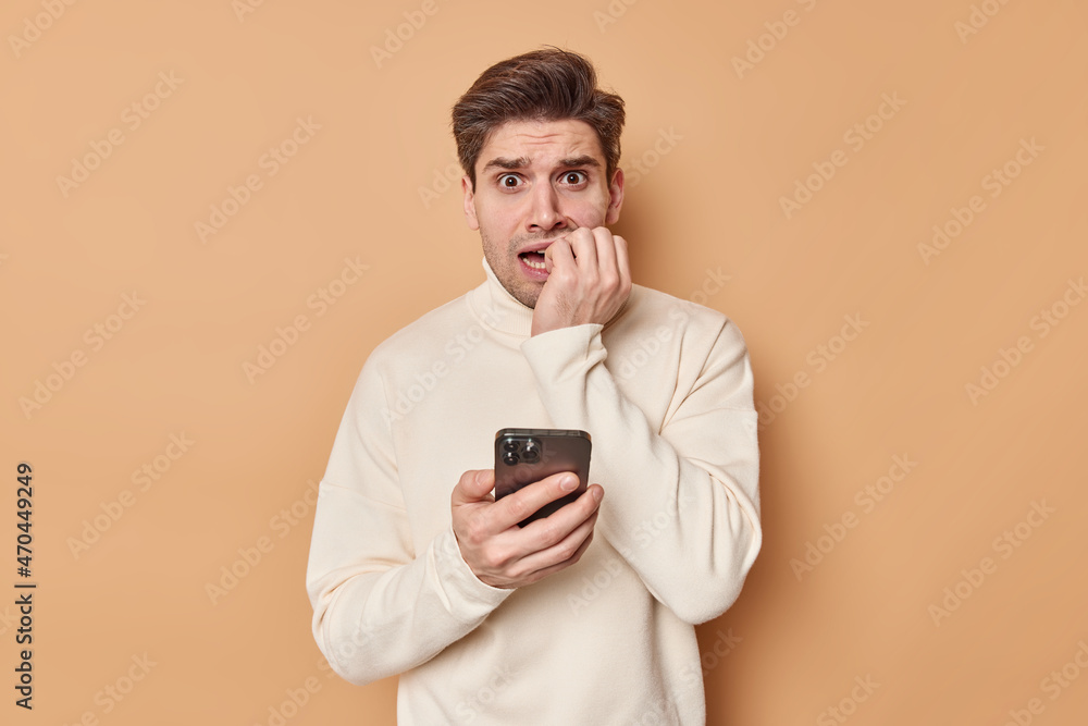 Anxious adult man bites finger nails reads chat nervously holds mobile phone wears casual jumper has scared expression made huge mistake isolated over beige background. What should I do help Anxious adult man bites finger nails reads chat nervously holds mobile phone wears casual jumper has scared expression made huge mistake isolated over beige background. What should I do help