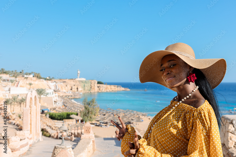Happy African American woman in yellow dress and sun hat enjoys view of coast of Red Sea on natural background. Panoramic views of blue sea with yachts and coastline, Sharm El Sheikh, Egypt. 