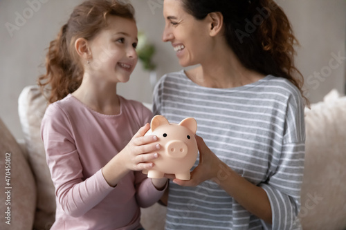 Close up happy mother with smiling little daughter holding pink piggy bank sitting on couch at home, caring mom and adorable girl child saving money for future, family insurance and investment
