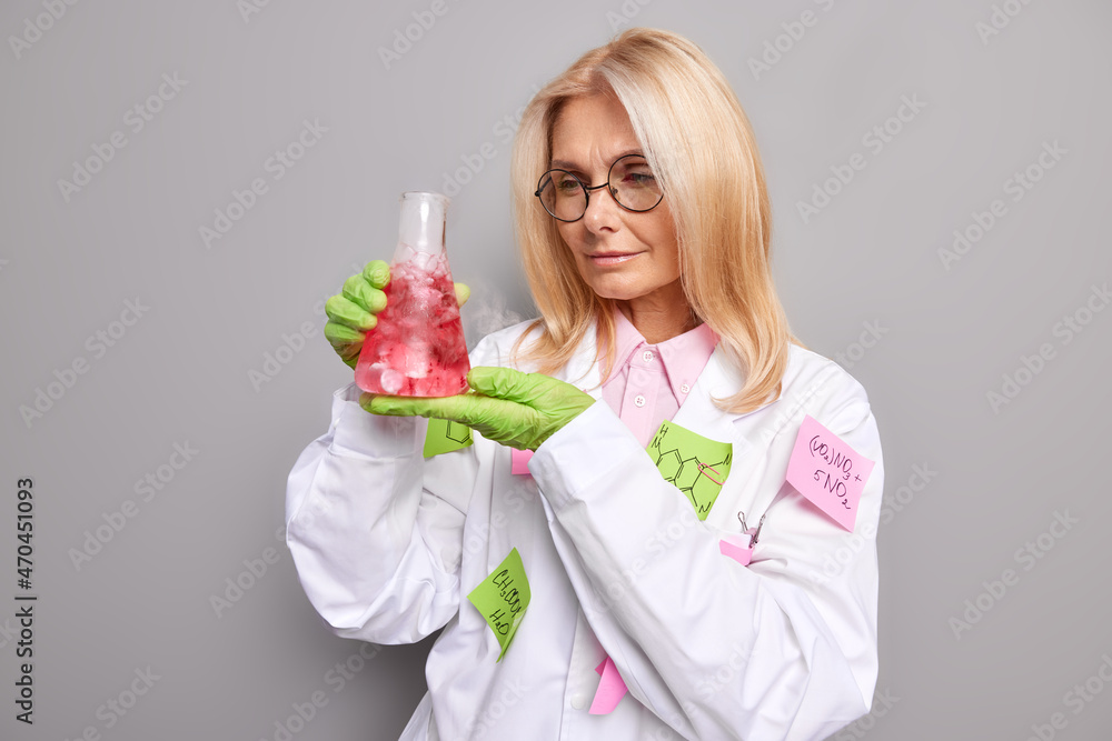 Female laboratory worker observes chemical processes focused at glass flask with red liquid wears medical coat and round spectacles isolated over grey background. Woman researcher conducts experiment Female laboratory worker observes chemical processes focused at glass flask with red liquid wears medical coat and round spectacles isolated over grey background. Woman researcher conducts experiment