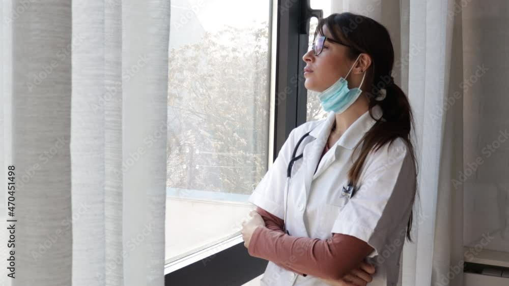 Young sad woman doctor taking a break standing and looking through the window wearing medical protective mask at work feeling tired and depressed after days of overworking shift at the hospital Young sad woman doctor taking a break standing and looking through the window wearing medical protective mask at work feeling tired and depressed after days of overworking shift at the hospital