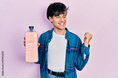 Handsome hipster young man holding detergent bottle screaming proud, celebrating victory and success very excited with raised arm