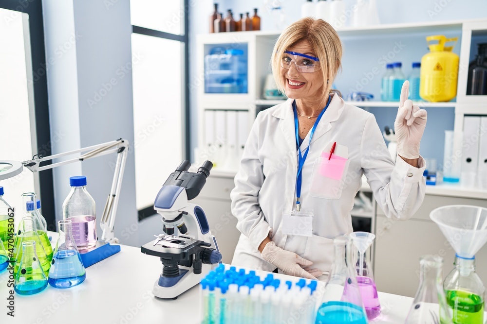 Middle age blonde woman working at scientist laboratory showing and pointing up with finger number one while smiling confident and happy. Middle age blonde woman working at scientist laboratory showing and pointing up with finger number one while smiling confident and happy.