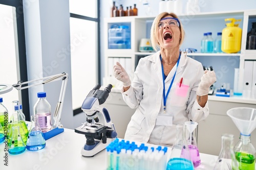 Middle age blonde woman working at scientist laboratory angry and mad screaming frustrated and furious  shouting with anger. rage and aggressive concept.