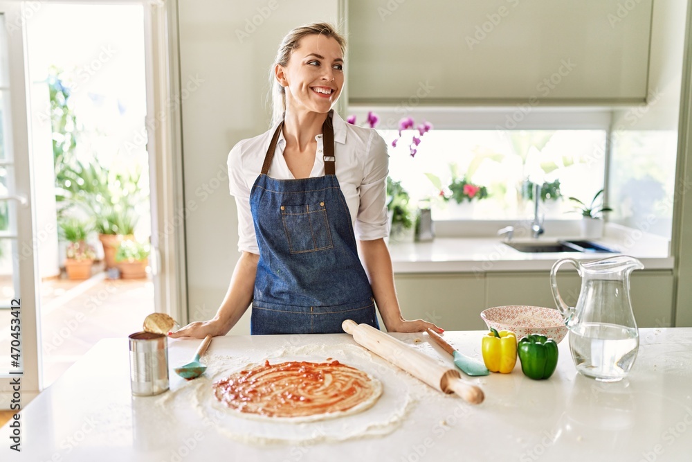 Beautiful blonde woman wearing apron cooking pizza looking away to side with smile on face, natural expression. laughing confident.