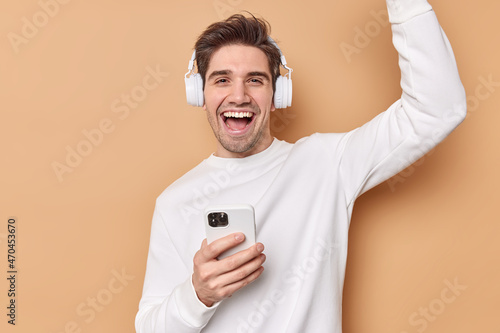 Glad cheerful man dances and laughs happily enjoys favorite playlist listens music via headphones holds mobile phone feels joyful isolated over beige background boosts mood with favorite song